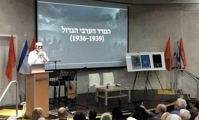מיטשניק פותר תעלומה של 130 שנה. מרצה לפני קהל. עומד על במה עם דוכן נואמים ומיקרופון ביד. על מסך מאחוריו מוקרן הכיתוב: המרד הערבי הגדול (1936-1939)