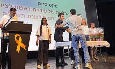 כוח משמעותי בעיר. מתתיהו והמלגאים. על במה איתי עומד מאחורי שולחן עם עוד אנשים. על השולחן עציצים עם פרחים. איתי לוחץ יד לצעיר העומד מולו מעבר לשולחן.